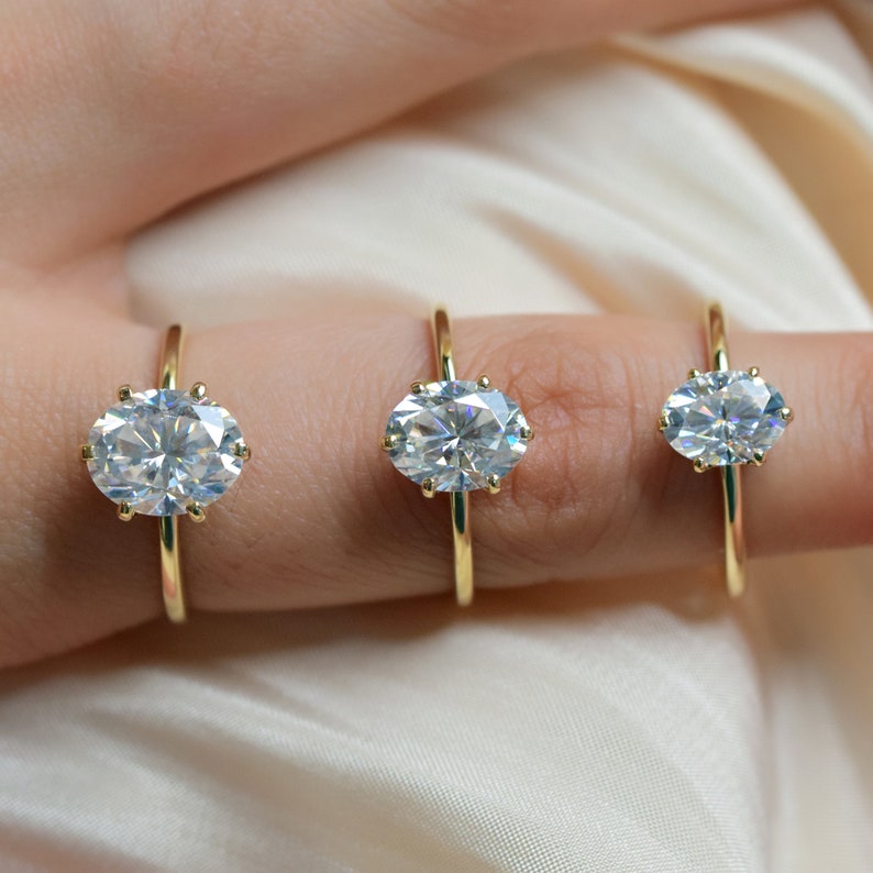 May include: Three gold rings with oval-shaped clear gemstones. The rings are on a finger and are different sizes.