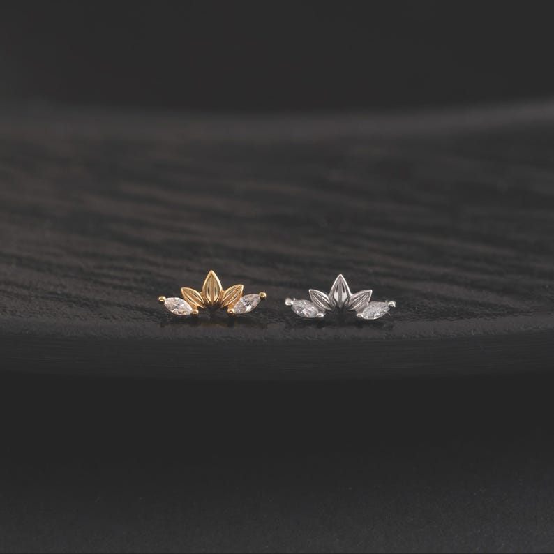 May include: Two small, decorative earrings. One is gold-toned, the other silver-toned. Each features a stylized floral design with three clear, marquise-cut gemstones. The earrings are set against a dark, textured background.
