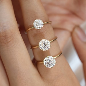 May include: Three gold engagement rings with round, clear diamonds. The rings are stacked on a finger, with the largest diamond on the bottom and the smallest diamond on top.