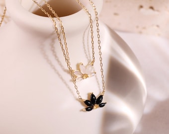 May include: Two delicate gold necklaces. The top necklace features a white flower pendant, while the bottom one showcases a black flower pendant. The necklaces are displayed against a soft, neutral background, highlighting the jewelry's elegance.