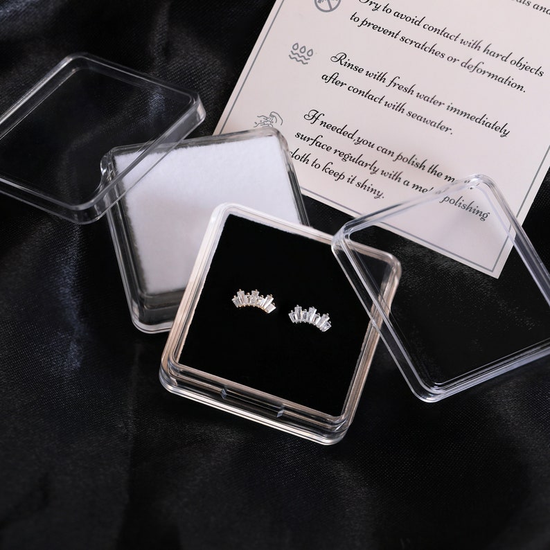 May include: A pair of silver-toned earrings in a clear plastic case with a black velvet interior. The earrings are shaped like a small crown and are displayed with a care instruction card. The case is open, revealing the earrings.