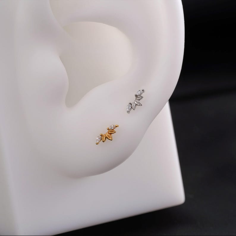 May include: Two ear studs, one gold-toned and one silver-toned, each featuring a cluster of small, leaf-shaped elements with clear, sparkling accents. The studs are displayed on a white ear model, showcasing their design and detail.