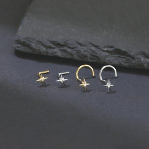 May include: A collection of four nose rings in gold and silver tones, each featuring a star-shaped design with a small, clear gemstone in the center. The nose rings are displayed on a dark, textured surface.