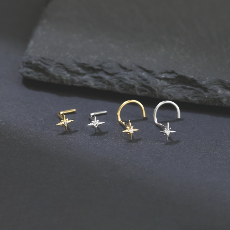 May include: A collection of four nose rings in gold and silver tones, each featuring a star-shaped design with a small, clear gemstone in the center. The nose rings are displayed on a dark, textured surface.