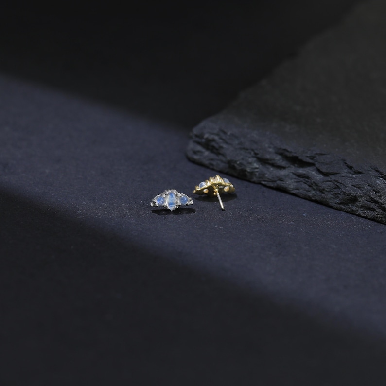 May include: A pair of gold-colored stud earrings, each featuring a fan-shaped design with three light blue gemstones. The earrings are displayed on a dark surface, with a natural stone element in the background.