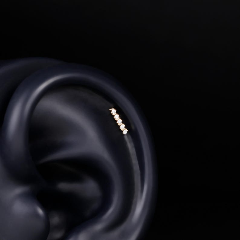 May include: A gold-toned ear piercing stud featuring a vertical row of five small, clear, round-cut stones set in a straight line. The jewelry is positioned on the upper cartilage area of a dark-colored, smooth, anatomical ear model.