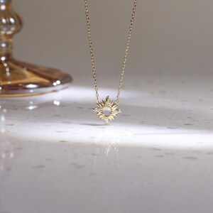 May include: A gold chain necklace featuring a sun-shaped pendant with a central round iridescent stone and small clear accent stones set within the rays. The pendant hangs from a delicate gold link chain, reflecting light on the surface below.