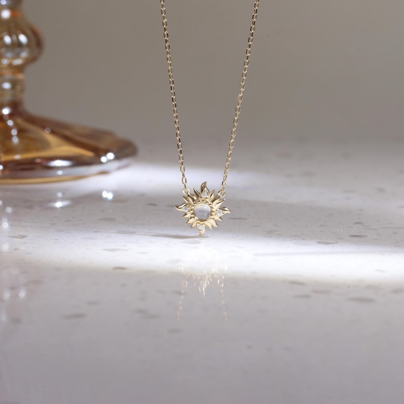 May include: A gold chain necklace featuring a sun-shaped pendant with a central round iridescent stone and small clear accent stones set within the rays. The pendant hangs from a delicate gold link chain, reflecting light on the surface below.