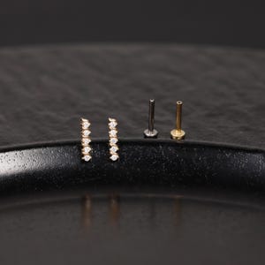 K&ouml;nnte beinhalten: Zwei goldfarbene Ohrstecker, die jeweils eine vertikale Reihe aus f&uuml;nf kleinen, klaren, rund geschliffenen Steinen aufweisen. Daneben liegen zwei flache Steckerstifte, einer in dunkler Metalloptik und einer in Goldoptik, die die R&uuml;ckseiten zeigen.