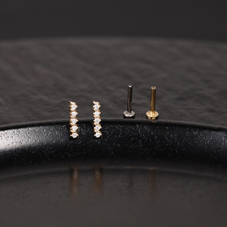 K&ouml;nnte beinhalten: Zwei goldfarbene Ohrstecker, die jeweils eine vertikale Reihe aus f&uuml;nf kleinen, klaren, rund geschliffenen Steinen aufweisen. Daneben liegen zwei flache Steckerstifte, einer in dunkler Metalloptik und einer in Goldoptik, die die R&uuml;ckseiten zeigen.