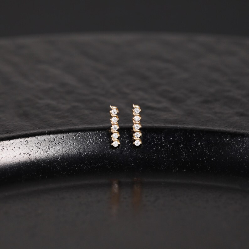 K&ouml;nnte beinhalten: Ein Paar Ohrstecker aus Gold, wobei jeder Ohrstecker ein vertikales Balkendesign mit f&uuml;nf kleinen, klaren, rund geschliffenen Steinen in einer linearen Reihe aufweist. Die Ohrringe haben eine polierte Metalloberfl&auml;che und einen sicheren Stiftverschluss.