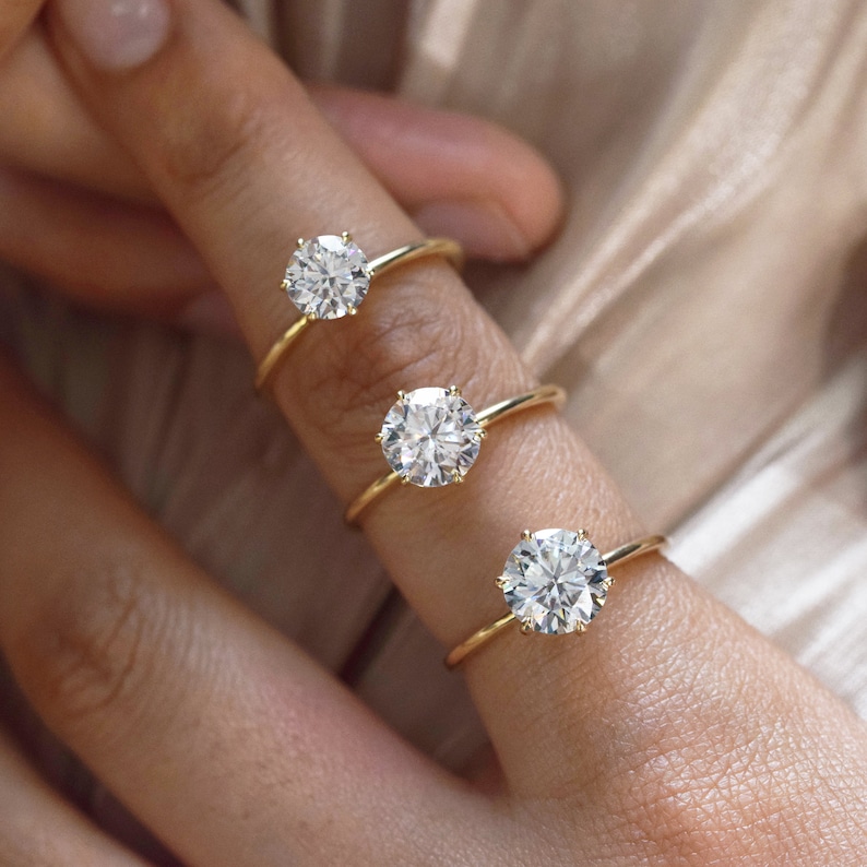 May include: Three gold engagement rings with different sized round diamonds. The rings are on a finger.