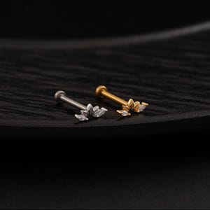 May include: Two pieces of jewelry, a silver and a gold labret stud, are displayed. Each stud features a floral design with clear, sparkling accents. The studs are set against a dark, textured background, highlighting their delicate details.