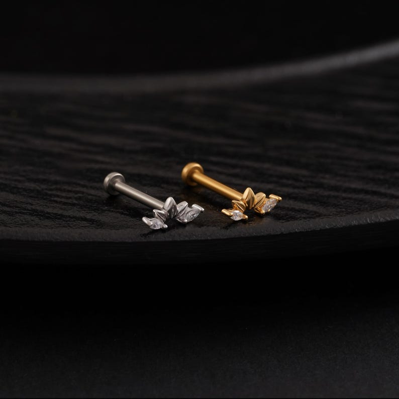 May include: Two pieces of jewelry, a silver and a gold labret stud, are displayed. Each stud features a floral design with clear, sparkling accents. The studs are set against a dark, textured background, highlighting their delicate details.