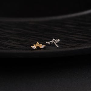 May include: Two small, decorative earrings. One is gold-toned with three clear, teardrop-shaped stones. The other is silver-toned with three similar stones. Both earrings have a floral design and are set against a dark background.