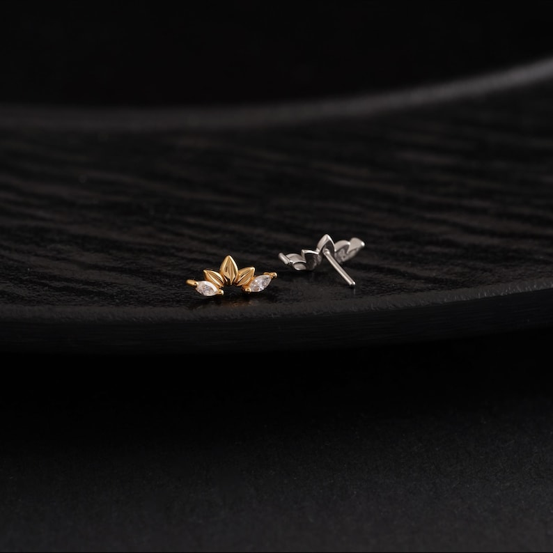 May include: Two small, decorative earrings. One is gold-toned with three clear, teardrop-shaped stones. The other is silver-toned with three similar stones. Both earrings have a floral design and are set against a dark background.
