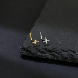 May include: Two nose studs, one gold and one silver, each featuring a four-pointed star design. The studs are set on a dark, textured surface, creating a contrast that highlights the jewelry's details. The star's points are adorned with small, sparkling accents.