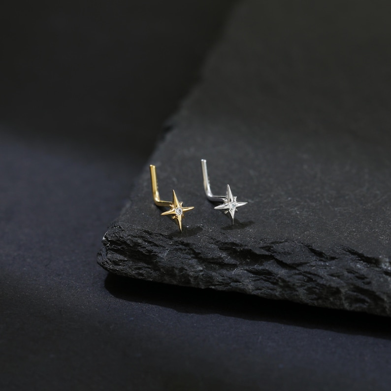 May include: Two nose studs, one gold and one silver, each featuring a four-pointed star design. The studs are set on a dark, textured surface, creating a contrast that highlights the jewelry's details. The star's points are adorned with small, sparkling accents.