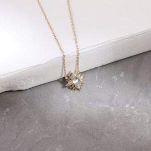 May include: A gold chain necklace featuring a sunburst-shaped pendant. The pendant has a central round iridescent moonstone surrounded by small clear stones set within radiating gold rays that create a star-like design on the metal frame.