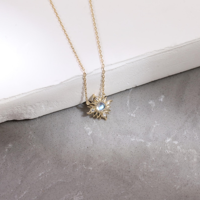 May include: A gold chain necklace featuring a sunburst-shaped pendant. The pendant has a central round iridescent moonstone surrounded by small clear stones set within radiating gold rays that create a star-like design on the metal frame.