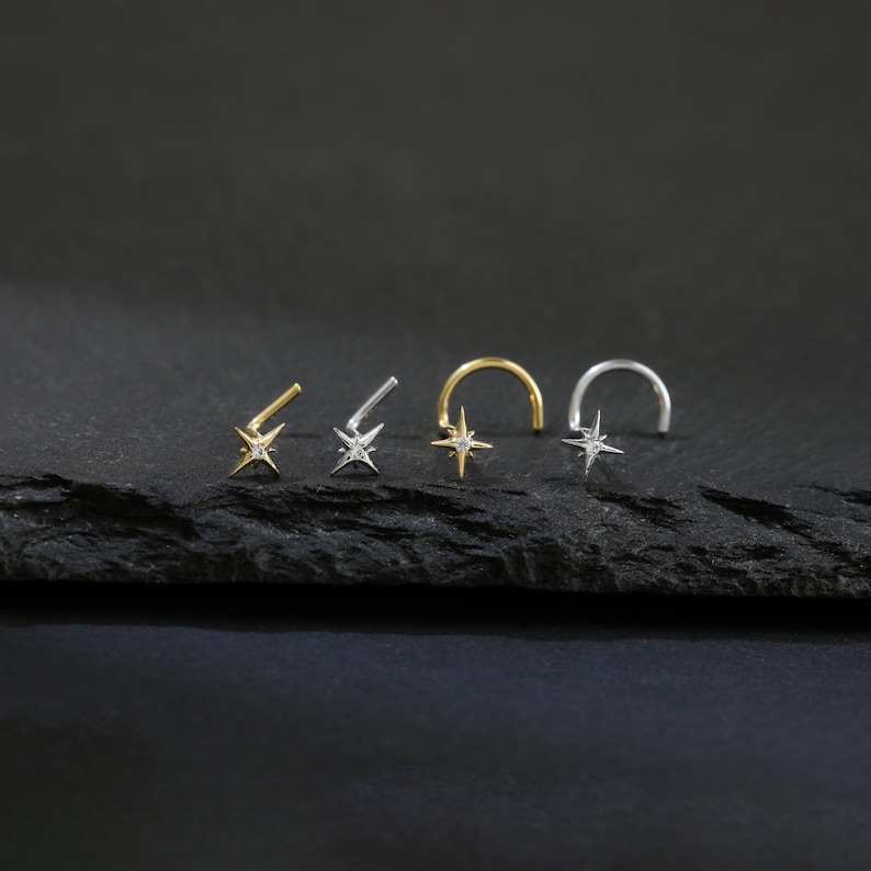 May include: Four nose rings with star-shaped designs. Two are straight, one is gold-toned, and the other is silver-toned. The other two are curved, one gold-toned and one silver-toned. Each star has a small, clear gem in the center.