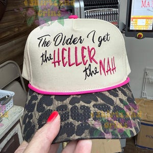 May include: A beige and leopard print baseball cap with the embroidered text "The Older I get the HELLER the NAH". The cap has a pink brim and trim. The cap is held by a person.
