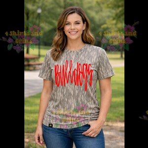 May include: A short-sleeved, crew neck t-shirt with a camouflage pattern in shades of gray and brown. The word "BULLDOGS" is written in red across the front. The person in the image is wearing blue jeans.