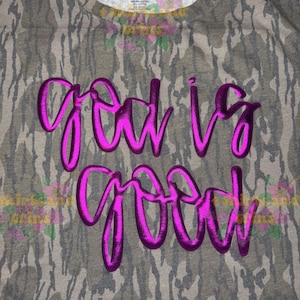 May include: A gray camouflage-patterned t-shirt with the phrase "God is Good" in bright pink, cursive lettering. The shirt has a crew neck and short sleeves. The fabric appears to be a blend of cotton and polyester.