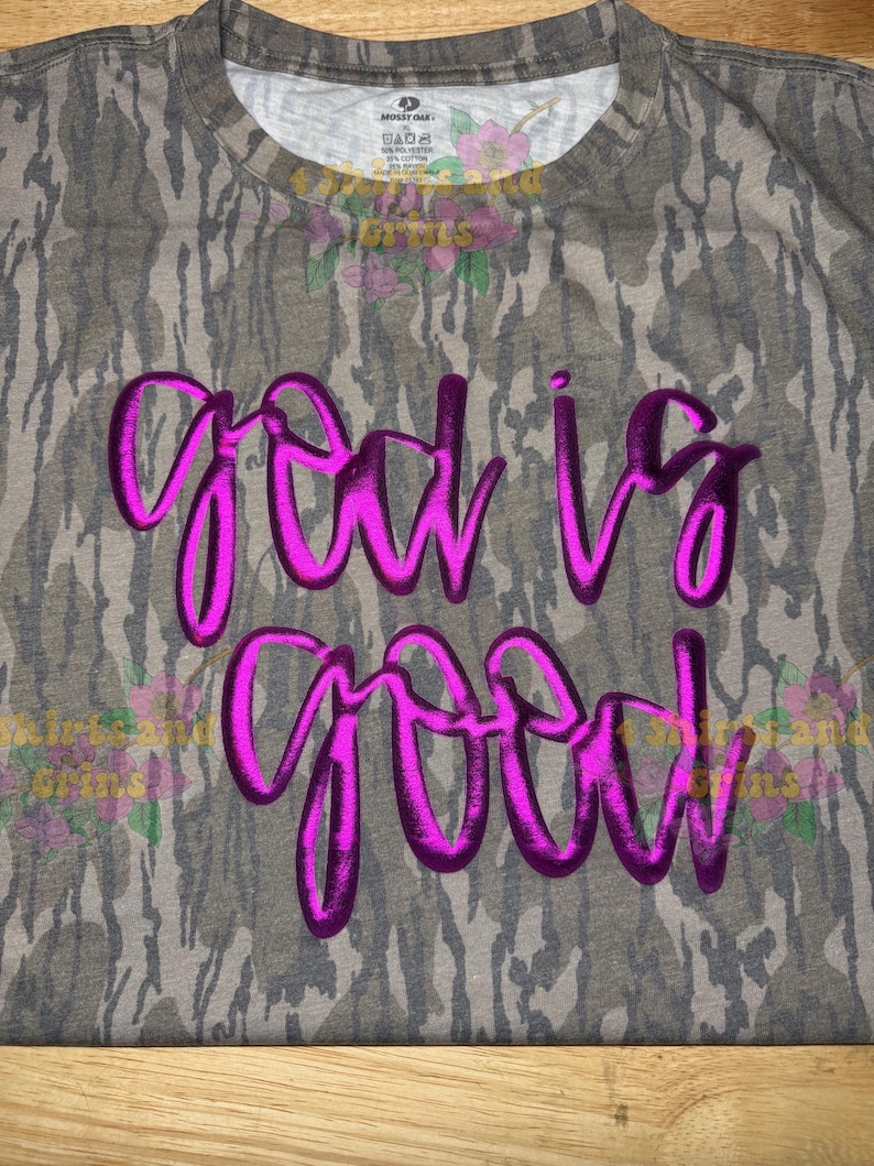 May include: A gray camouflage-patterned t-shirt with the phrase "God is Good" in bright pink, cursive lettering. The shirt has a crew neck and short sleeves. The fabric appears to be a blend of cotton and polyester.