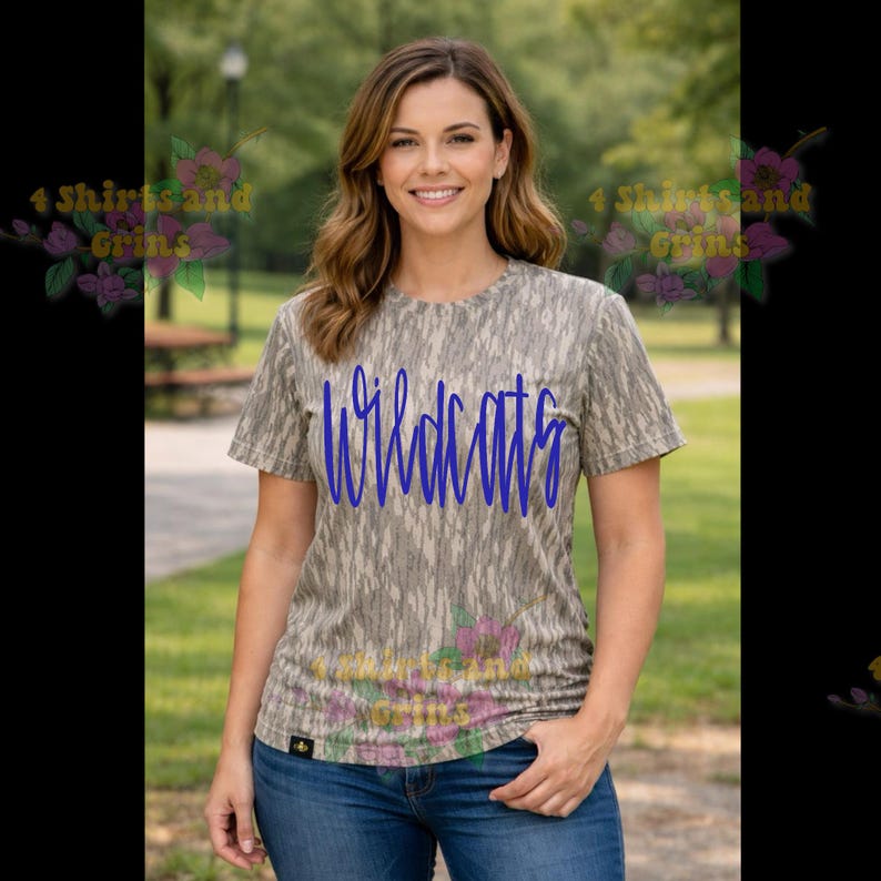 May include: A short-sleeved, camouflage-patterned t-shirt with the word "Wildcats" in blue script. The shirt is worn with blue jeans. The background is a park setting with trees and a walking path.