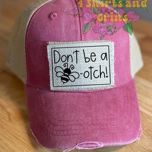 May include: Pink and beige trucker hat with a distressed look. The hat has a patch sewn on the front that says "Don't be a -otch!" with a bee illustration.
