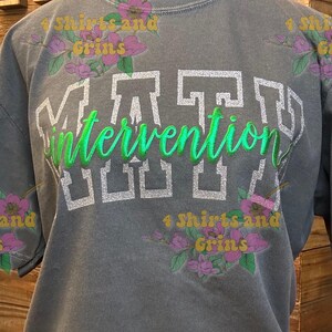 May include: Gray short-sleeved t-shirt with the word "MATT" in white block letters above the word "Intervention" in green cursive. The shirt has a vintage look and floral accents.