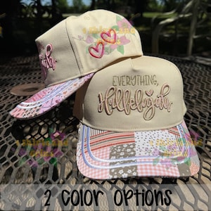 May include: Two beige baseball caps with embroidered designs. One cap features the word "Faith" in pink script, the other says "Everything, Hallelujah". Both have patterned brims with floral and patchwork designs. The image also says "2 color options".