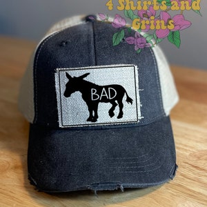 May include: A black and white distressed trucker hat with a white mesh back. The hat has a patch sewn on the front that says "BAD" with a silhouette of a donkey.