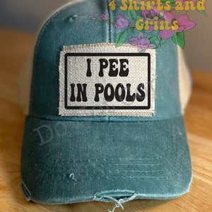May include: A green and beige trucker hat with a distressed look. The hat has a patch sewn on the front that says "I PEE IN POOLS".