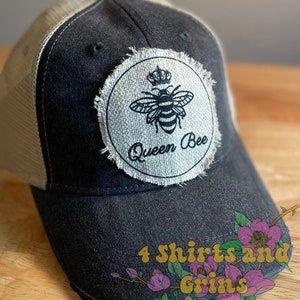 May include: Black and white trucker hat with a distressed, beige fabric patch that says "Queen Bee" and features a bee with a crown. The hat has a mesh back.