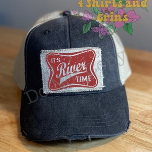 May include: A black and white trucker hat with a distressed look. The hat has a red and white patch with the text "It's River Time" on it.
