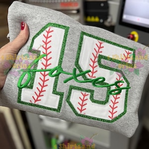 May include: A gray sweatshirt with the number "15" in a baseball design. The number is white with red stitching, outlined in green glitter. The word "Foxes" is embroidered in green script.