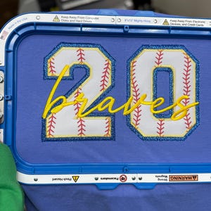 May include: A blue fabric with the number "20" and the word "Braves" embroidered in yellow. The numbers are white with red stitching and a blue and yellow border, resembling a baseball. The text is in a cursive font.