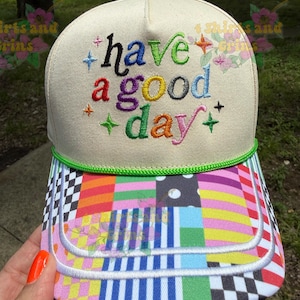 May include: A cream-colored baseball cap featuring the embroidered text have a good day in multi-colored letters with small star accents. The cap includes a bright green cord trim and a brim decorated with a vibrant, multi-patterned patchwork design.