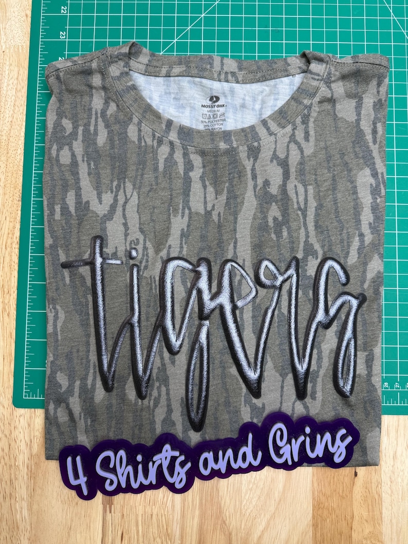 May include: A camouflage-patterned t-shirt with the word "Tigers" in a stylized, shadowed font. Below the shirt is a purple sticker that reads "4 Shirts and Grins". The shirt is folded on a green cutting mat.