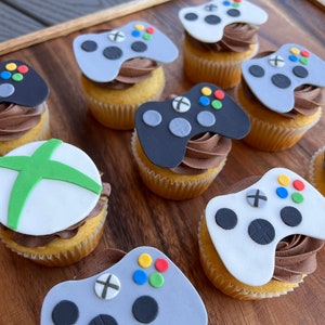 May include: A collection of cupcakes decorated with fondant game controllers. The controllers are in various colors, including black, white, and gray. Some of the controllers have green, red, blue, and yellow buttons.