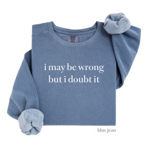 May include: A blue jean colored sweatshirt with the text "i may be wrong but i doubt it" in white. The sweatshirt has a crew neck and the sleeves are rolled up. The bottom right corner of the sweatshirt has the words "blue jean".