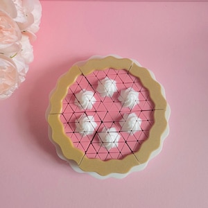 Op de afbeelding: Een ronde speelgoedtaart met een beige geschulpte korst en een roze vulling met een geometrisch driehoekig patroon. Zes witte toefjes slagroom zijn in een cirkel op de roze bovenkant gerangschikt, rustend op een witte ondergrond.