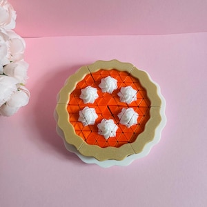 Op de afbeelding: Een rond taartvormig puzzelspeelgoed met een feloranje oppervlak dat bestaat uit kleine driehoekige segmenten. De puzzel is omlijst door een beige, geschulpte korstrand en is afgewerkt met vijf witte, stervormige toefjes slagroom.