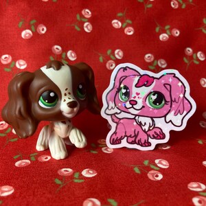 May include: Two small plastic toy dogs. The dog on the left is brown and white with brown eyes. The dog on the right is pink with green eyes and glitter. Both dogs are on a red background with white flowers.