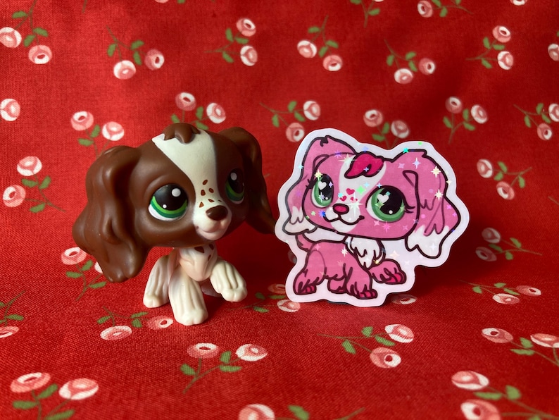 May include: Two small plastic toy dogs. The dog on the left is brown and white with brown eyes. The dog on the right is pink with green eyes and glitter. Both dogs are on a red background with white flowers.