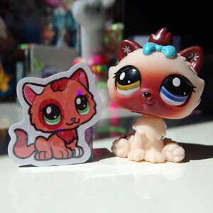 May include: A red and white cartoon cat sticker with green eyes and a pink nose. The sticker is on a white background. A brown and white toy cat with blue eyes and a pink bow is sitting on a white surface.