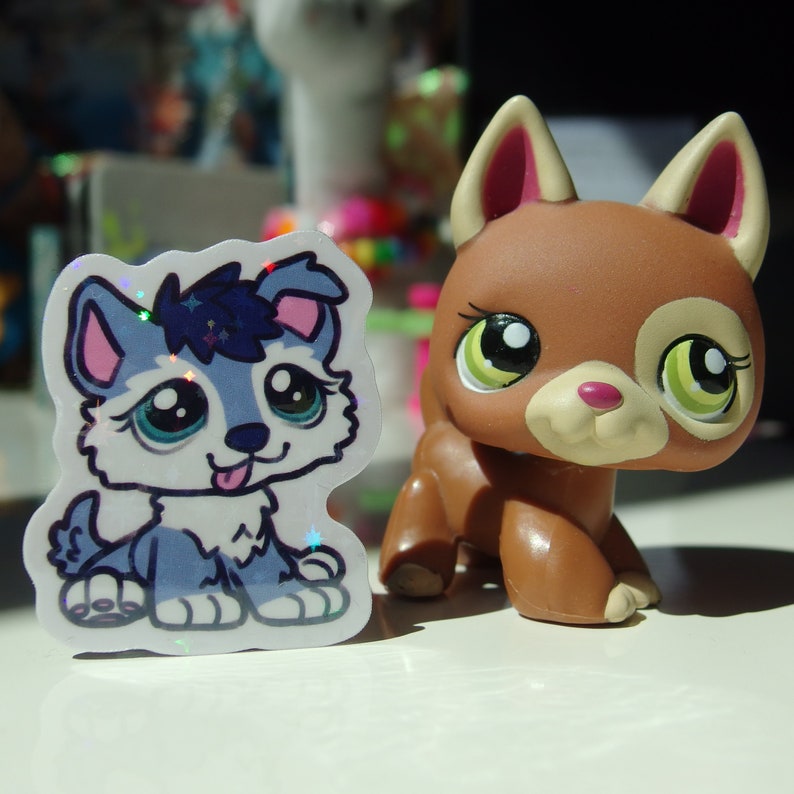 May include: A sticker of a blue and white husky puppy with a pink nose and tongue sticking out. The sticker is next to a brown and white plastic toy dog with green eyes.
