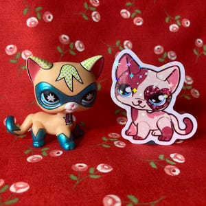 May include: Two toy cats, one with a superhero mask and the other with a pink heart on its eye, are on a red background with a floral pattern.